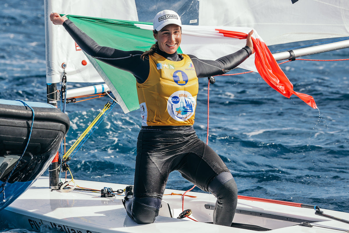An ILCA sailor in the yellow leader's bib kneeling on her boat, holding her national flag above her head